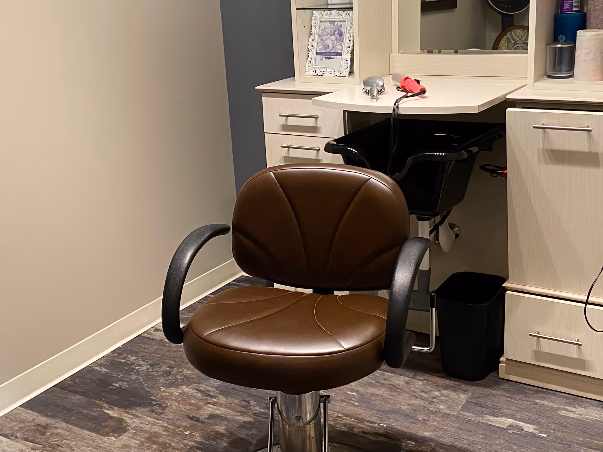 A salon chair with brown cushioned seat and backrest, black armrests, positioned in front of a hair washing station with a black basin and a countertop holding a hair dryer and other hair care items.
