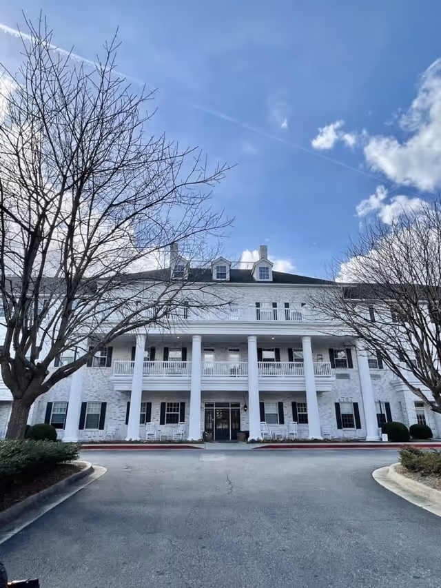 Front exterior view of a large white senior living facility building with multiple windows, black shutters, and a covered porch supported by white columns. There are leafless trees on either side of the driveway leading up to the entrance under a blue sky with some clouds.