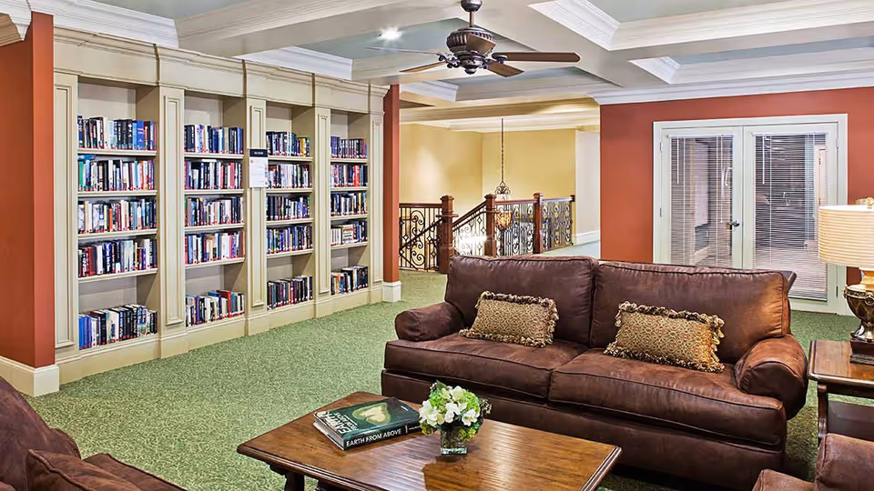 A cozy living room area in a senior living facility featuring a brown leather sofa with decorative pillows, a wooden coffee table with a book and a small flower arrangement, built-in bookshelves filled with books, a ceiling fan, and a lamp on a side table. The room has green carpet flooring and warm-colored walls with white trim.