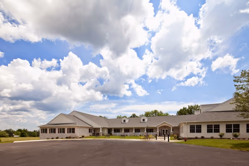 Exterior view of a single-story retirement community building under a partly cloudy sky. The building has a light-colored facade with multiple windows and a covered entrance. There is a paved driveway and some landscaping with grass and small shrubs around the building.