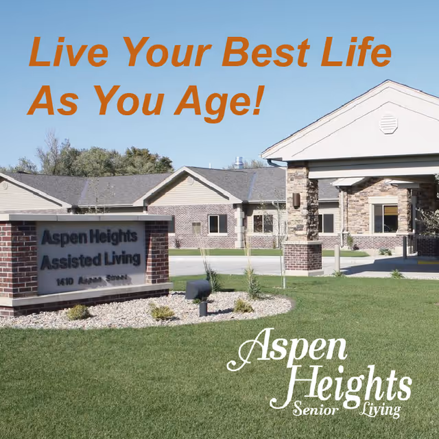 Exterior view of Aspen Heights Assisted Living facility showing a brick sign with the facility name and address, a well-maintained lawn, and the building entrance under a clear blue sky.