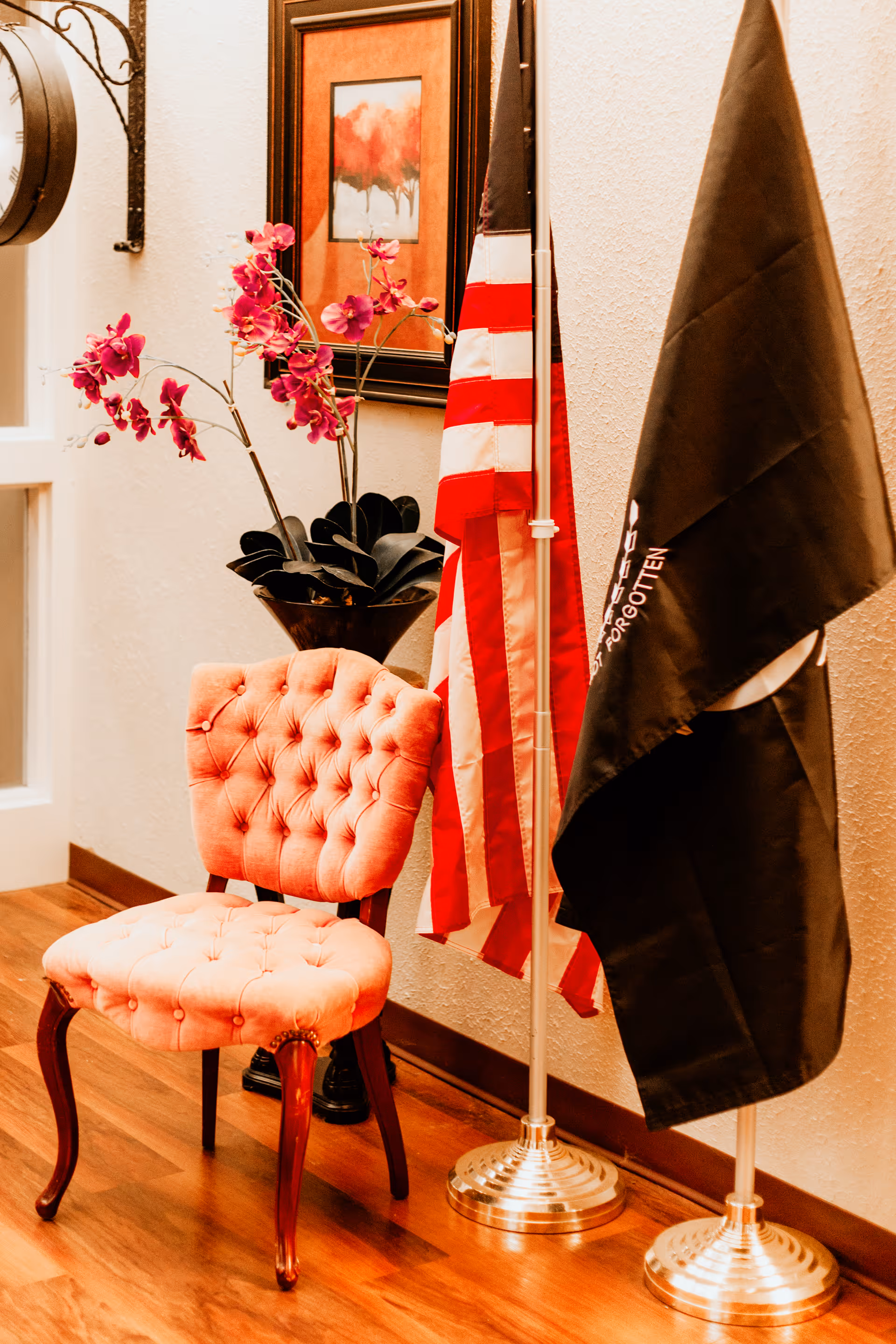 A cozy corner with a pink tufted chair on a wooden floor, next to two flags on silver stands, one American flag and one black flag. Behind the chair is a black vase with pink flowers and a framed painting of red trees on the wall.