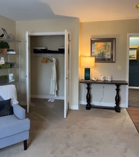 A cozy interior room featuring an open closet with a white robe and slippers inside. To the right of the closet is a dark wooden console table with a blue lamp, two mugs, and a small decorative item. Above the table hangs a framed picture. On the left side, part of a gray upholstered chair with a white throw blanket and a dark pillow is visible. The room has beige carpet and light-colored walls.