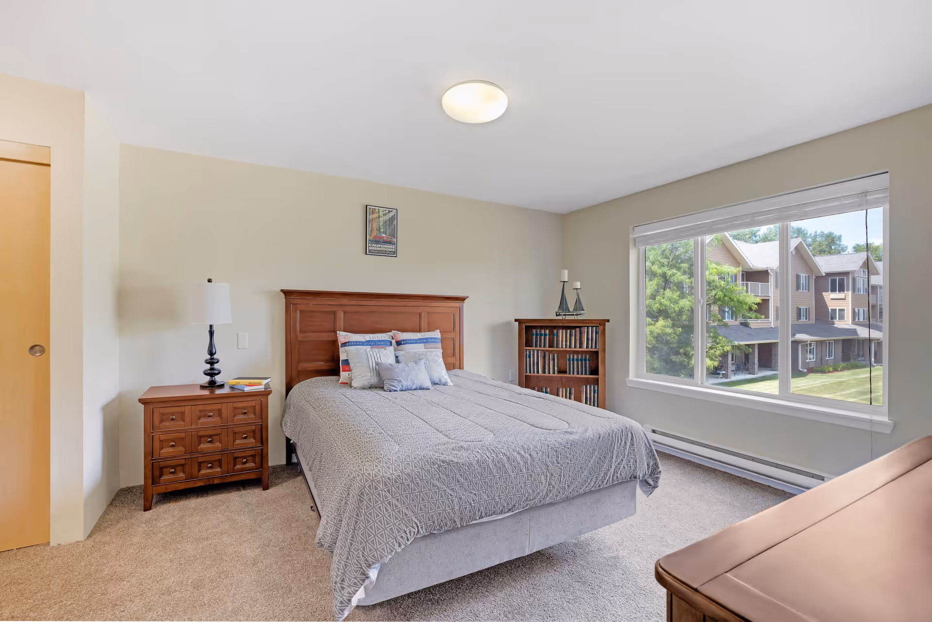 A bright bedroom with a large window showing an outside view of a residential building. The room features a wooden bed with a gray bedspread and several pillows, a wooden nightstand with a lamp and books, a wooden bookshelf with candles on top, and beige carpeted floor.