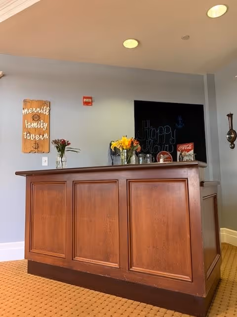 A wooden bar counter with paneling in a room with light gray walls and a beige carpet. On the counter are vases with flowers and some small decorative items. Behind the counter, a blackboard with 'Happy Hour' written on it is visible. A wooden sign on the wall reads 'merrell family tavern'.