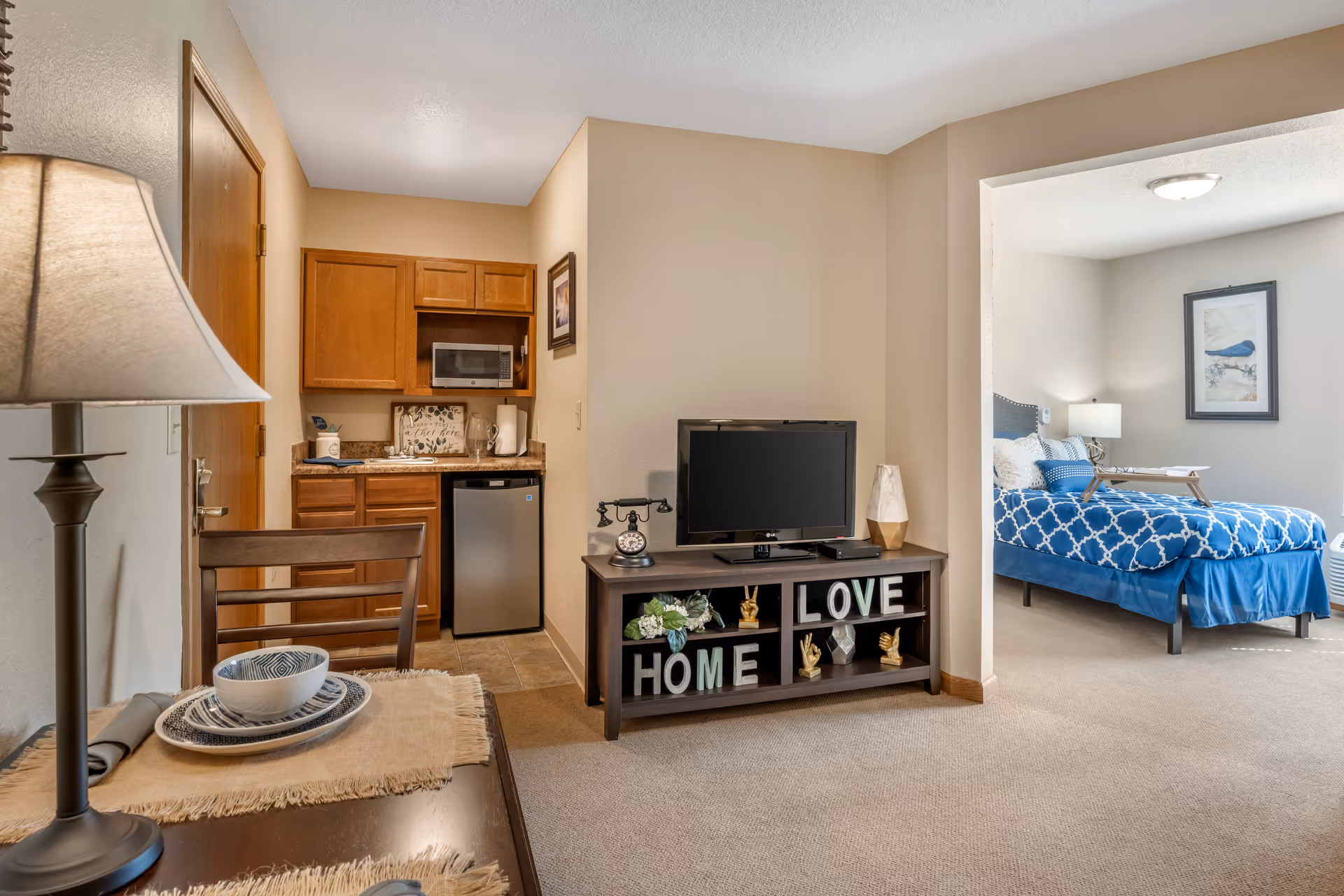 Interior view of a senior living facility apartment at Brookdale Greenville featuring a small kitchenette with wooden cabinets, a microwave, and a mini refrigerator. Adjacent to the kitchenette is a dark wooden TV stand with a flat-screen TV and decorative items spelling out 'HOME' and 'LOVE'. To the right, an open doorway leads to a bedroom with a bed dressed in blue and white patterned bedding, a bedside lamp, and framed artwork on the wall. A table with a lamp and place setting is visible in the foreground.