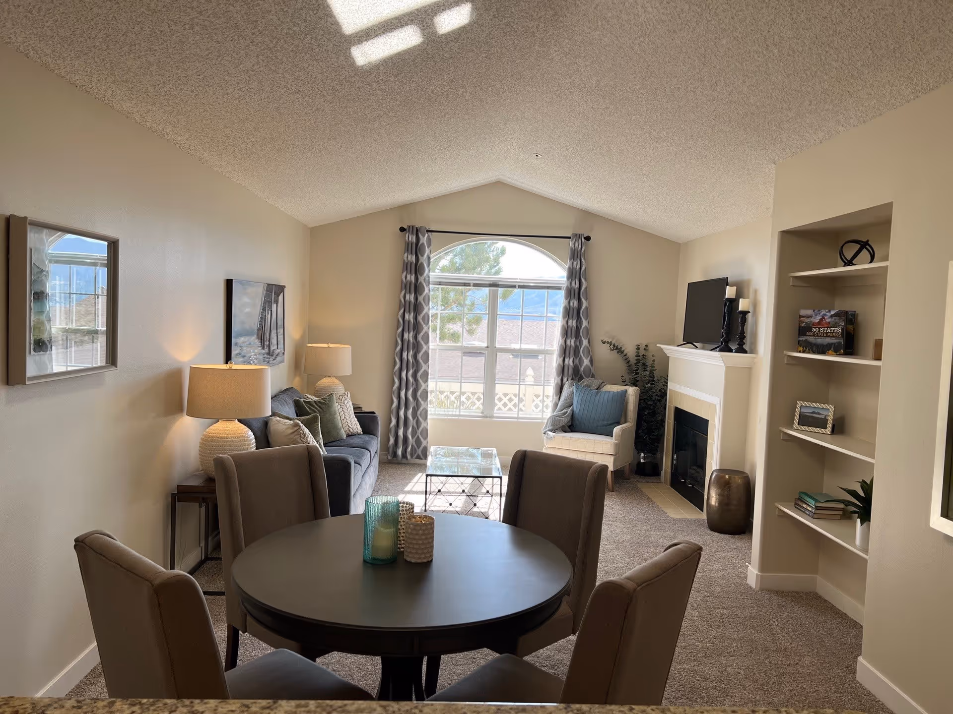 A cozy living room with a round dining table and four chairs in the foreground. The room features a gray sofa with multiple cushions, a glass coffee table, a beige armchair with a blue pillow, and a fireplace with a TV on the mantel. There are built-in shelves with books and decorative items on the right side. A large window with patterned curtains lets in natural light, and the walls are painted a light beige color.