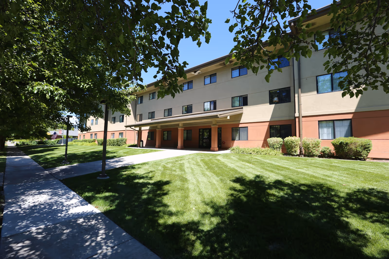 Three-story senior living building with a covered entrance, manicured lawn, trees, and a sidewalk in front.