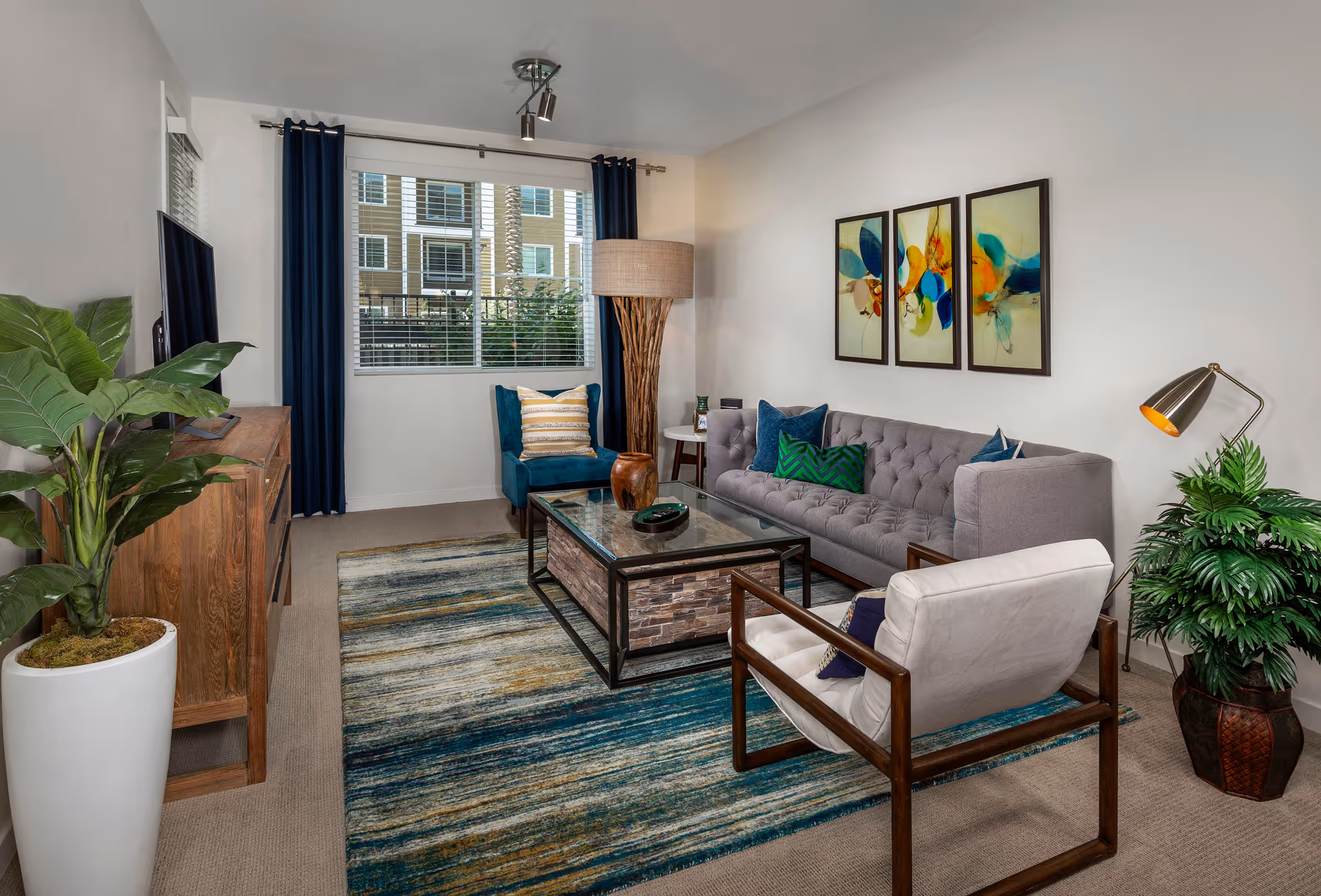 A modern living room with a gray tufted sofa adorned with blue and green pillows, a white cushioned armchair with wooden arms, and a blue accent chair with a striped pillow. A rectangular glass coffee table with a stone base sits on a multicolored blue and beige rug. The room features a wooden media console with a flat-screen TV, two large potted plants, a tall floor lamp with a woven shade, and three abstract colorful paintings on the white wall. A window with white blinds and dark blue curtains lets in natural light.