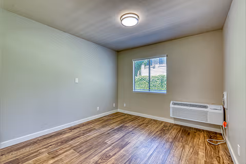 Empty room with wooden flooring, beige walls, a window with blinds, a ceiling light fixture, and a wall-mounted air conditioning unit.