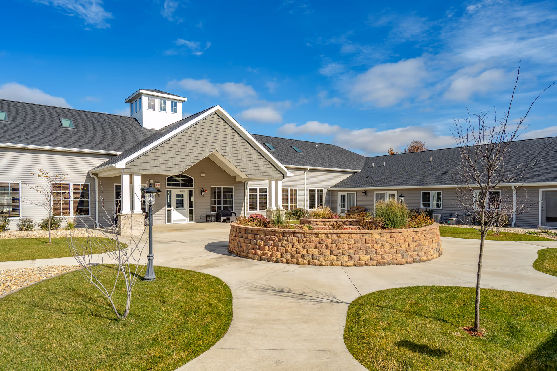 Exterior view of Villas of Holly Brook Assisted Living & Memory Care in Pekin, IL, showing a single-story building with a covered entrance, a circular brick planter with plants, a concrete walkway, and a well-maintained lawn under a partly cloudy blue sky.