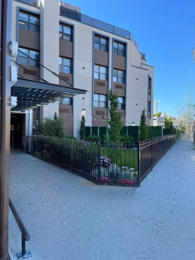 Three-story beige and brown building with a fenced landscaped front yard, entrance canopy, and sidewalk.