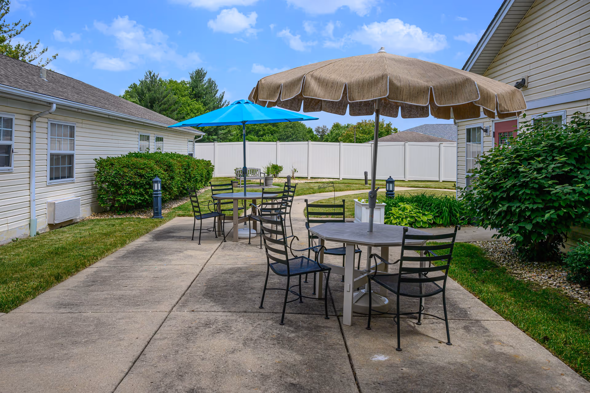 Outdoor patio area with round tables and black metal chairs, each table shaded by large umbrellas, one beige and one blue. The patio is surrounded by green bushes and grass, with white fencing and light-colored buildings in the background under a partly cloudy blue sky.
