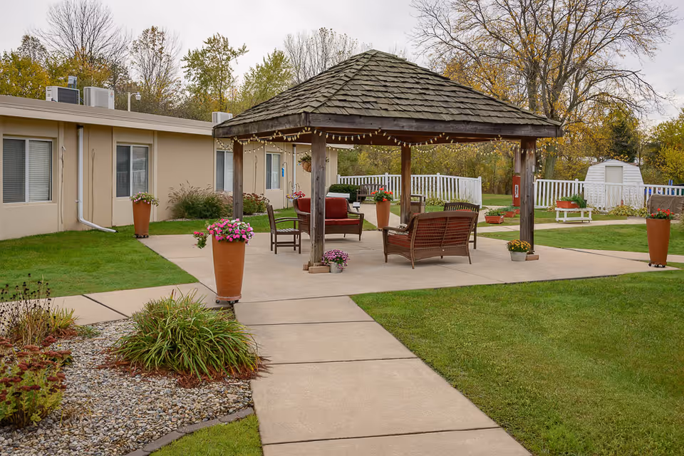 Landscaped outdoor courtyard featuring a wooden gazebo with seating, potted flowers, and paved walkways beside a single-story building.