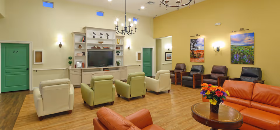 Communal living room with rows of armchairs and sofas facing a TV cabinet, colorful wall art, and a small round table with flowers.