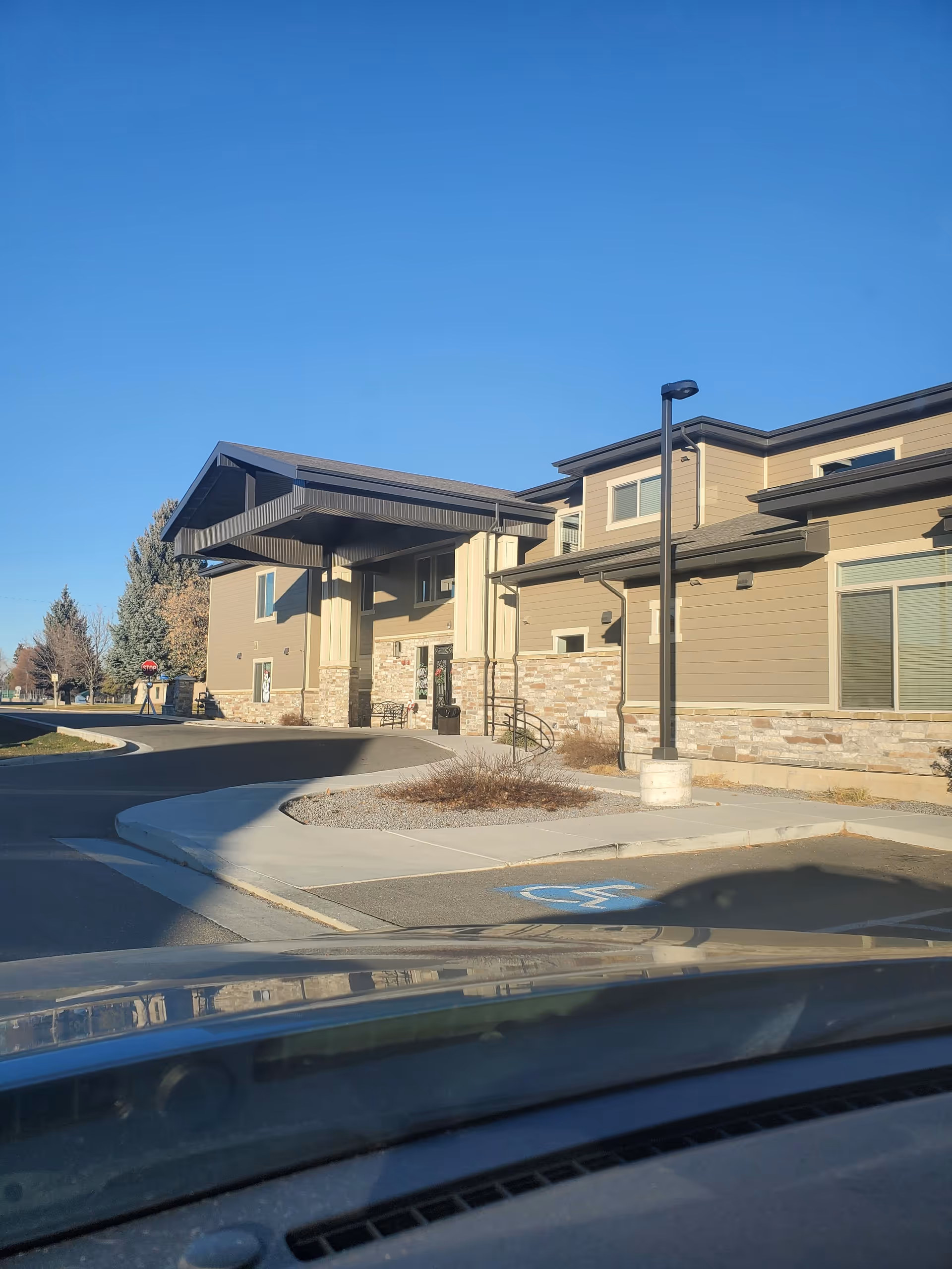 Exterior view of Blacksmith Fork Assisted Living facility showing the front entrance with a covered drop-off area, beige siding with stone accents, a sidewalk, a handicap parking space, and a clear blue sky.