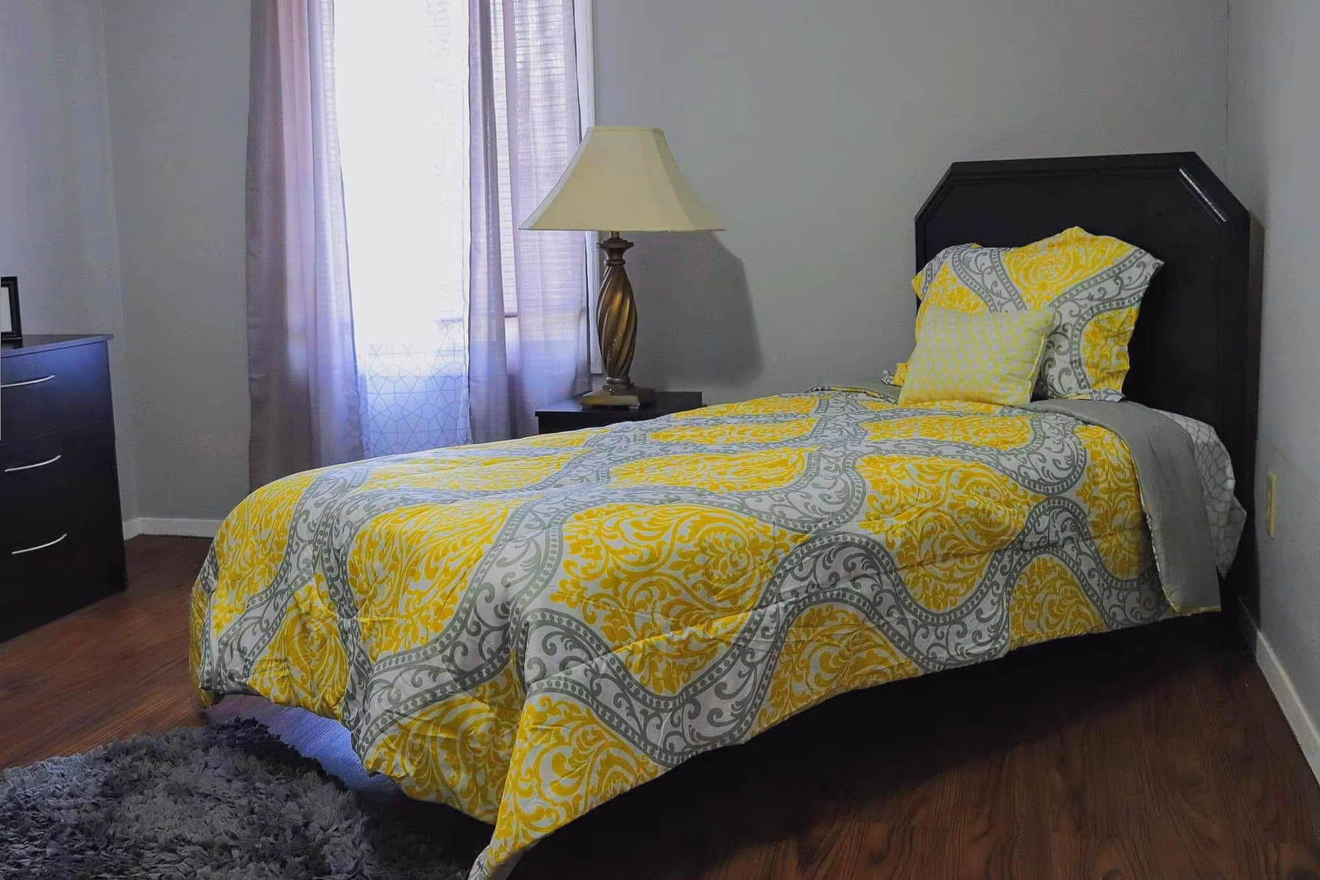 A bedroom with a single bed featuring a yellow and gray patterned comforter and matching pillows. There is a wooden nightstand with a lamp beside the bed, a window with sheer curtains letting in natural light, a dark dresser on the left, and a gray rug on the wooden floor.