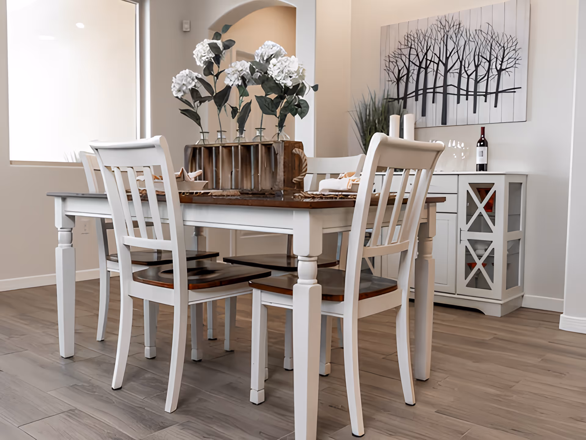 Bright dining room with a white wooden table and chairs, floral centerpiece and a sideboard beneath tree wall art.