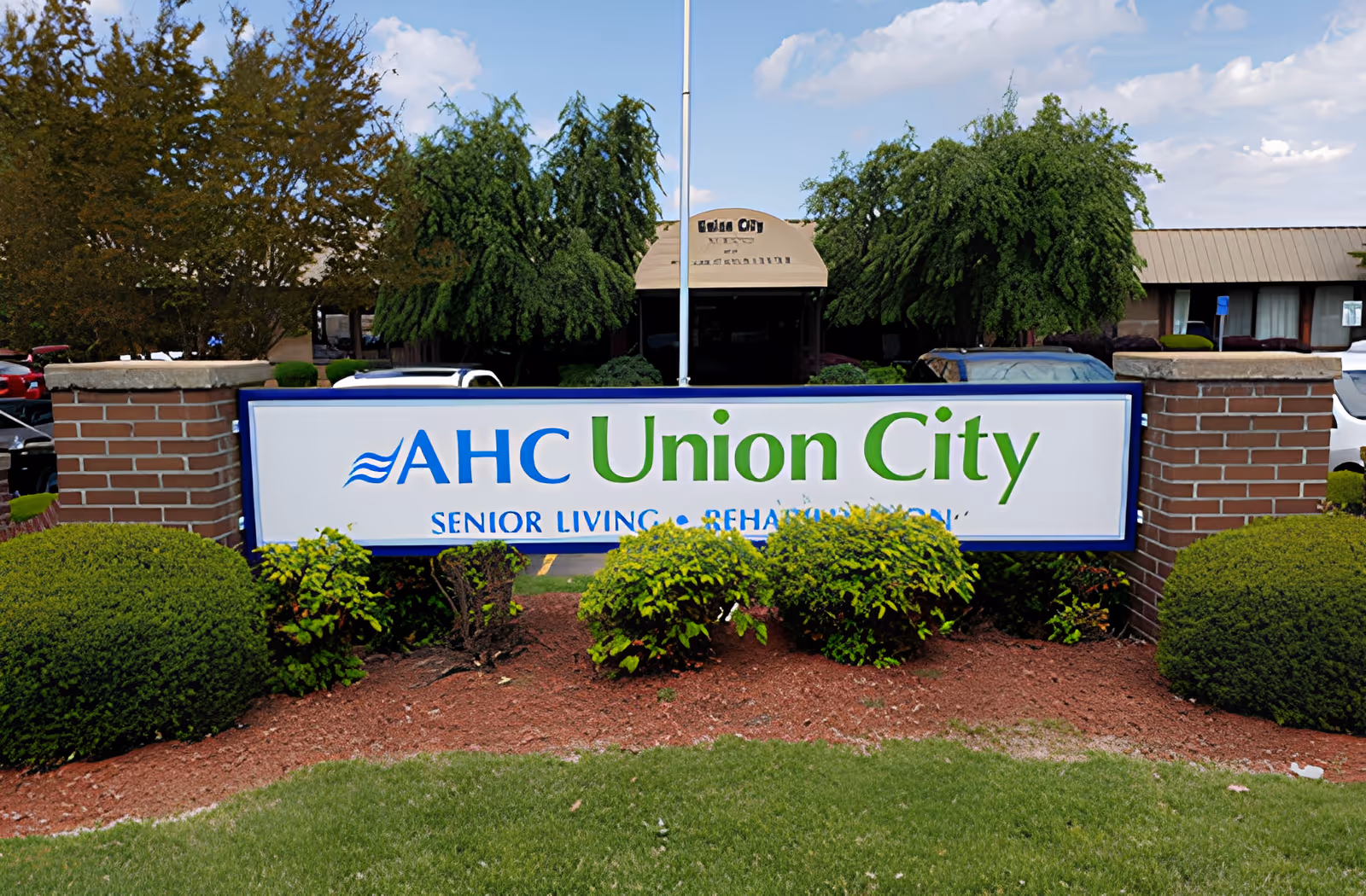 Outdoor view of the entrance sign for AHC Union City senior living facility, surrounded by green bushes and trees with a building and parked cars in the background under a partly cloudy sky.