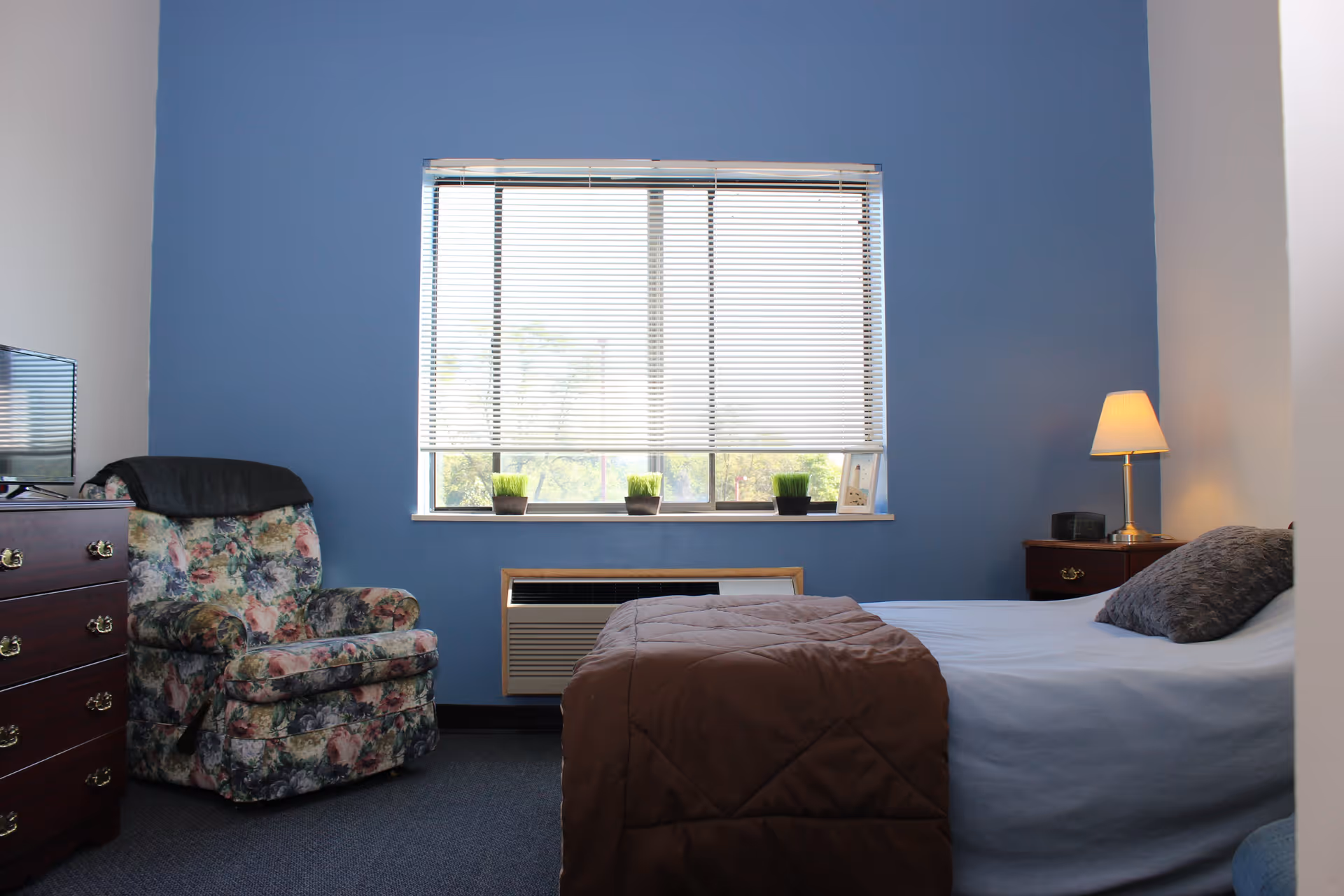 A small bedroom with a bed covered in a light blue sheet and brown blanket, a floral upholstered armchair, a wooden dresser with a TV on top, a window with white blinds partially open, and a nightstand with a lamp and clock.