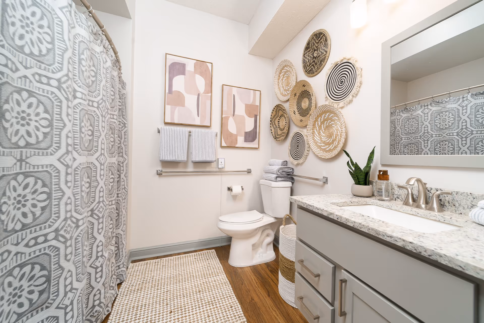 A modern bathroom featuring a white toilet, a gray and white patterned shower curtain, a granite countertop with a sink, and a large mirror. The walls are decorated with abstract art and woven circular wall hangings. There are two gray hand towels hanging on a rack above the toilet, and a small plant and toiletries on the countertop. The floor has a wooden finish with a beige and white patterned rug.
