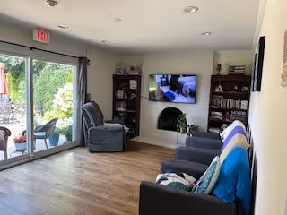 A cozy living room with wooden flooring, a dark sofa with colorful blankets, a recliner chair, a wall-mounted TV, bookshelves, and a sliding glass door leading to an outdoor patio area with greenery.