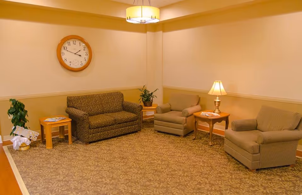 A cozy sitting area with a patterned loveseat, two armchairs, two wooden side tables with a lamp and magazines, a potted plant, a wall clock showing 3:45, and warm beige walls with a carpeted floor.