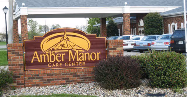 Brick entrance sign reading 'Amber Manor Care Center' in front of the facility with shrubs and parked cars.