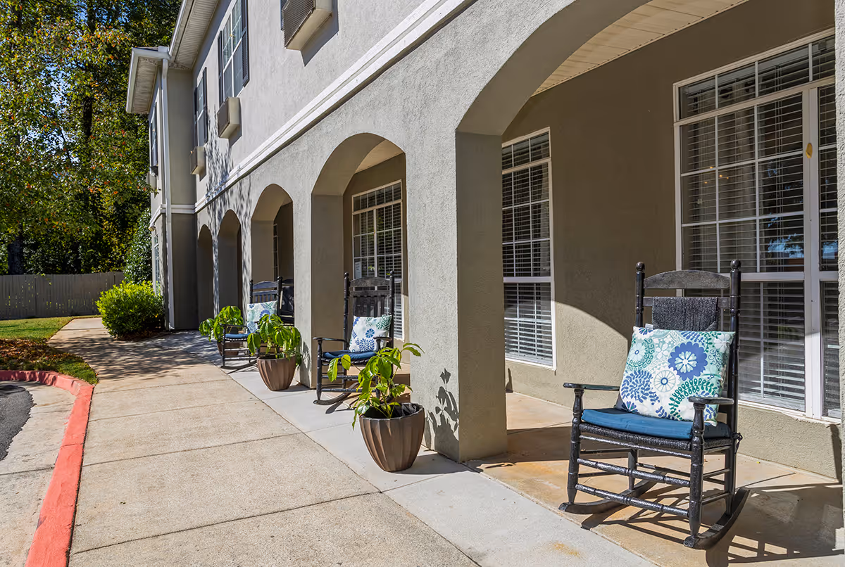 Outdoor covered patio area at Greenwood Place Assisted Living and Memory Care with black rocking chairs featuring blue and white patterned cushions, potted plants, and large windows with white blinds. The building exterior is light gray with arches and a concrete walkway beside a red-painted curb.