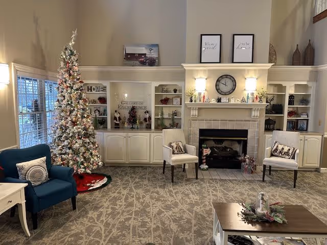 A cozy living room decorated for Christmas with a tall, white and pink decorated Christmas tree near large windows. There are two white armchairs with patterned pillows placed on either side of a fireplace with a clock and framed quotes above it. Built-in shelves flank the fireplace, displaying various decorative items. A blue armchair and a small white side table are positioned near the tree. A coffee table with holiday decorations is in the foreground on a patterned carpet.