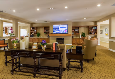 A cozy living room area in Manzanita Village featuring a large flat-screen TV mounted above a fireplace, built-in wooden shelves with books and decorative plants, comfortable armchairs, and a wooden table with candles, flowers, and framed photos. The room is warmly lit with recessed ceiling lights and has a patterned carpet.