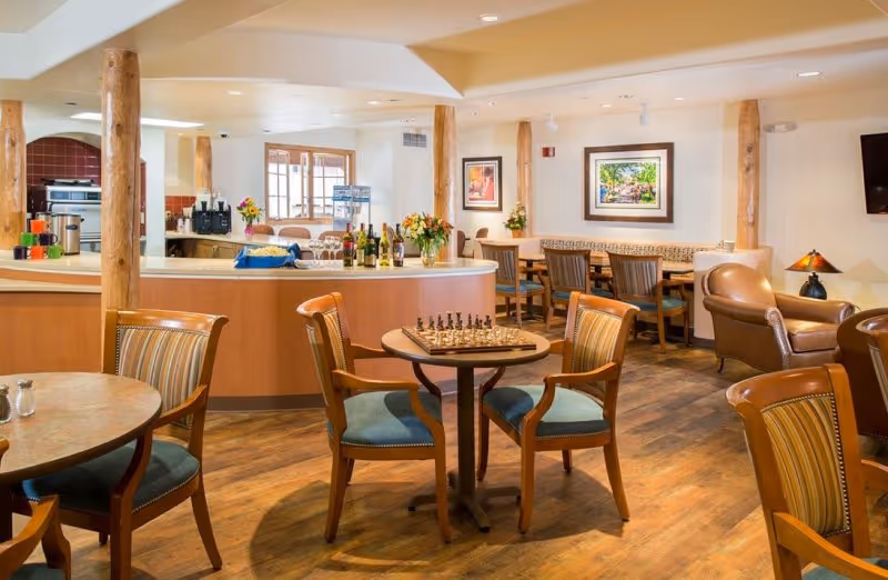 A cozy common area with wooden floors and furniture, featuring several tables and chairs. One table has a chessboard set up. There is a counter with bottles and flowers, and a kitchen area in the background. The walls are decorated with framed artwork, and there is a television mounted on the right wall.