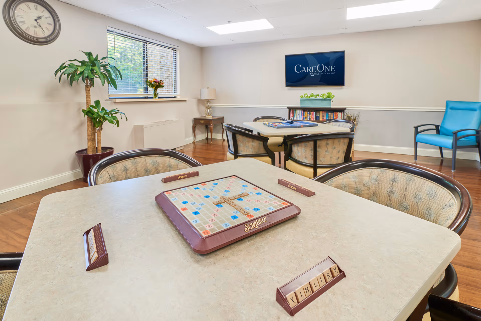 A well-lit common room with a Scrabble game set up on a table in the foreground. The room has several cushioned chairs around tables, a large wall clock, a potted plant, a window with blinds, a small side table with a lamp, a bookshelf with books, and a wall-mounted TV displaying the CareOne logo.