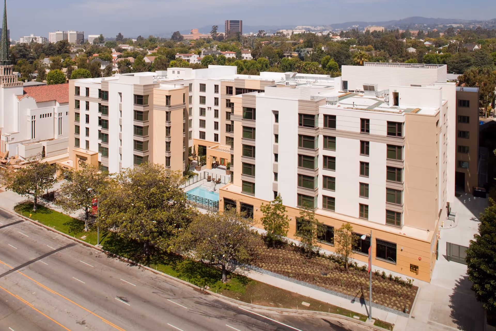 Aerial view of Belmont Village Senior Living Westwood, a multi-story senior living facility with beige and white exterior walls, surrounded by trees and landscaping. The building features multiple windows and a small outdoor pool area. The facility is located along a wide street with several lanes and sidewalks.