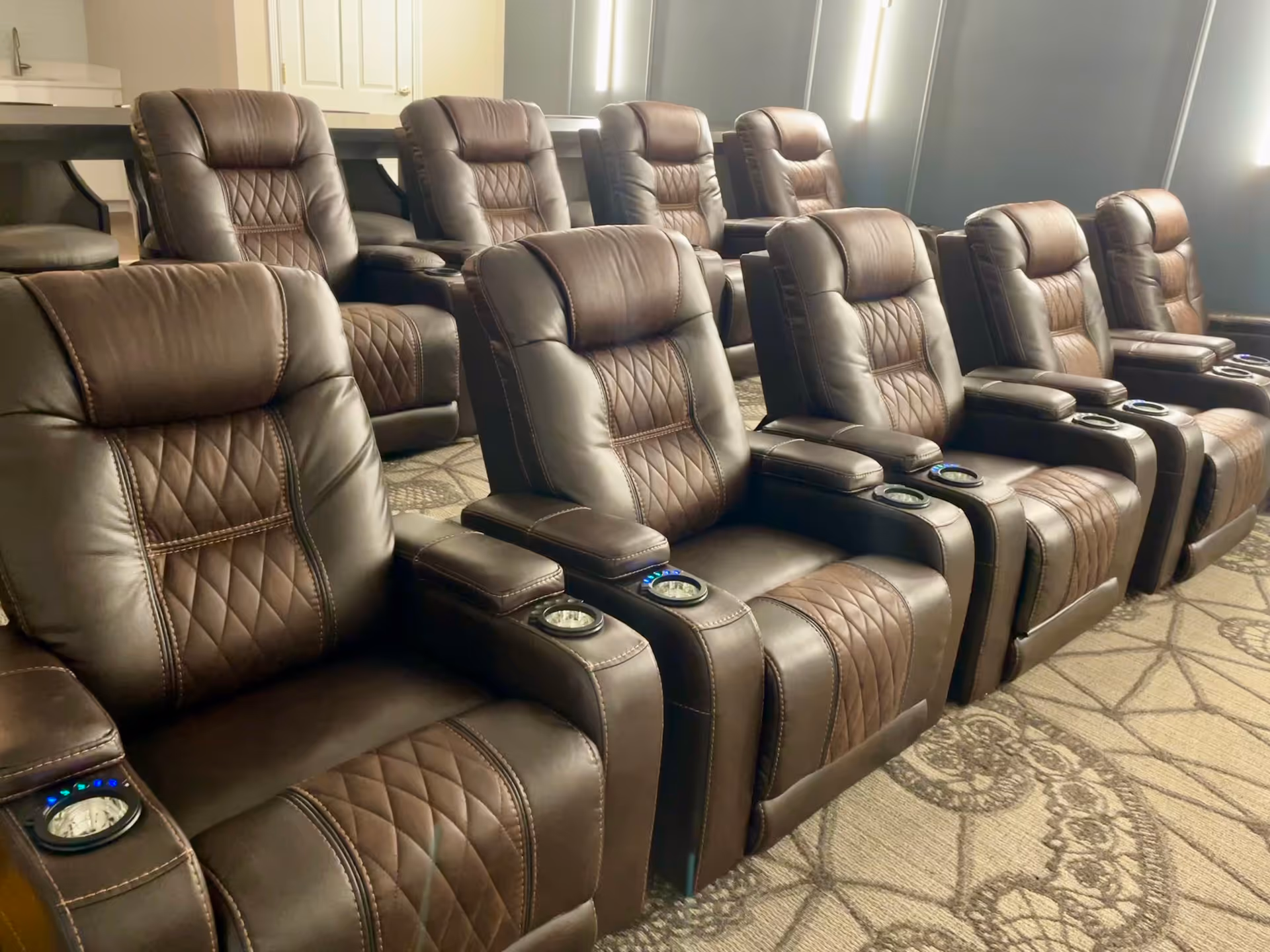 A row of brown leather recliner chairs with cup holders in a room with patterned carpet and soft lighting, likely a small theater or media room.