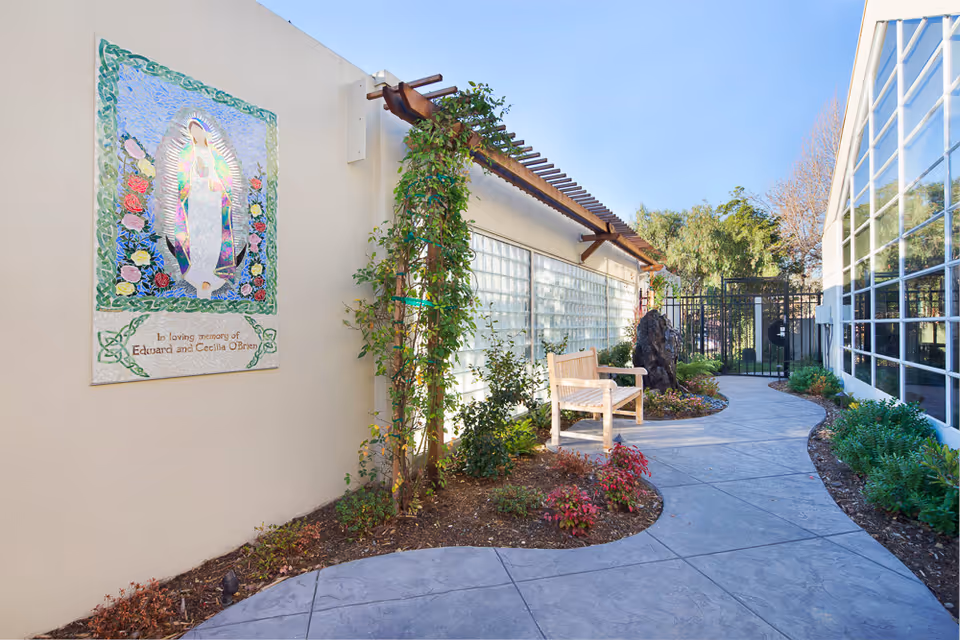 A peaceful outdoor garden path at Mercy Retirement & Care Center featuring a curved concrete walkway, a wooden bench, climbing plants on a trellis, various shrubs and flowers, a decorative mosaic plaque on the wall, and a black metal gate at the end of the path.