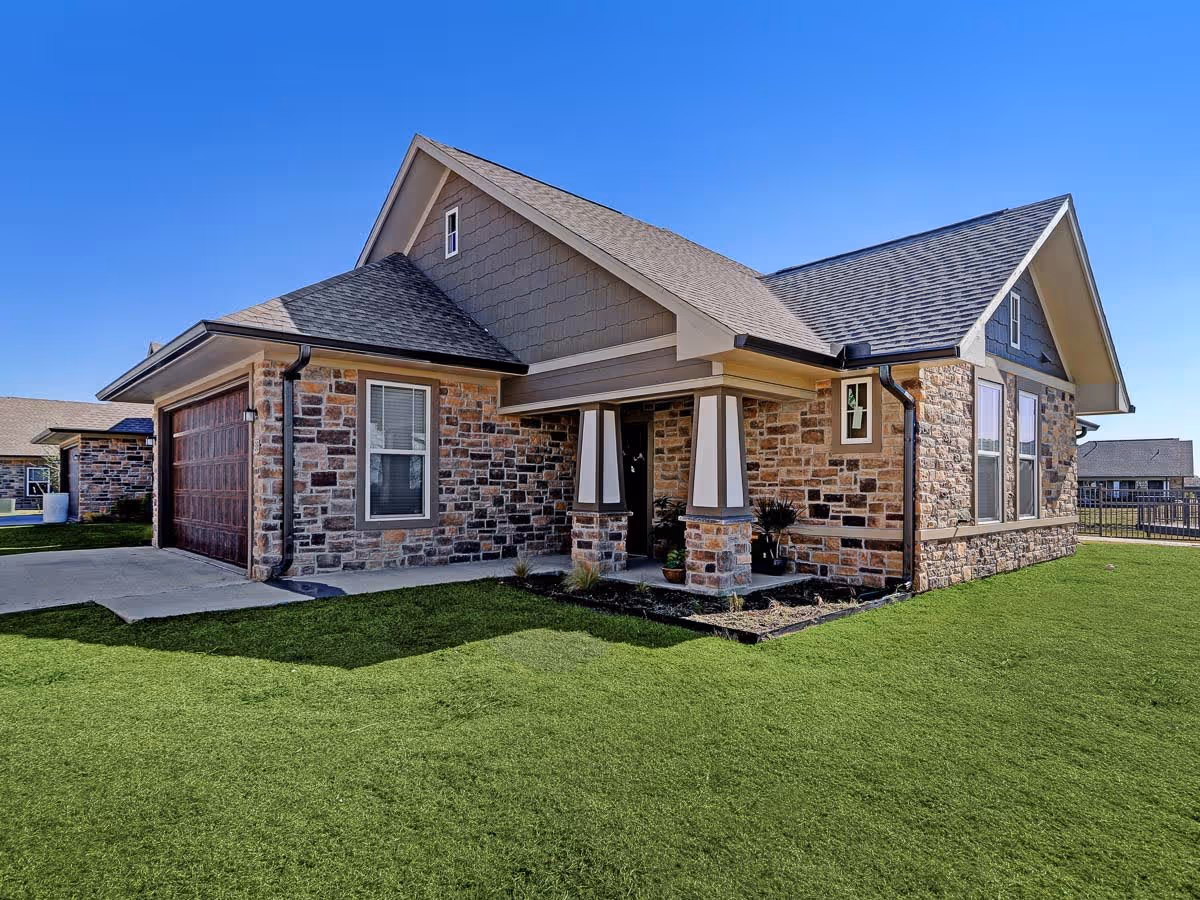 Exterior view of a single-story stone house with a gabled roof, a garage with wooden doors, and a small covered porch supported by two columns. The house is surrounded by a well-maintained green lawn under a clear blue sky.