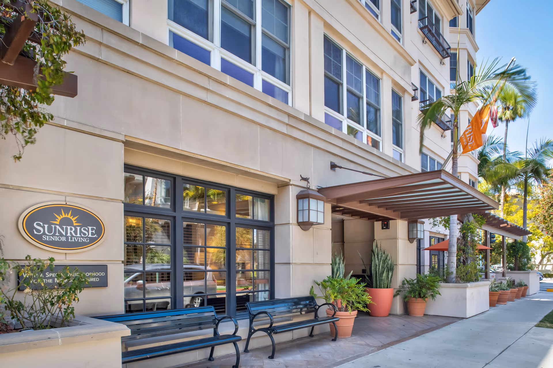 Exterior view of Sunrise Senior Living facility in Beverly Hills showing the building facade with large windows, two black benches, several potted plants, and an orange Sunrise flag under a covered entrance.