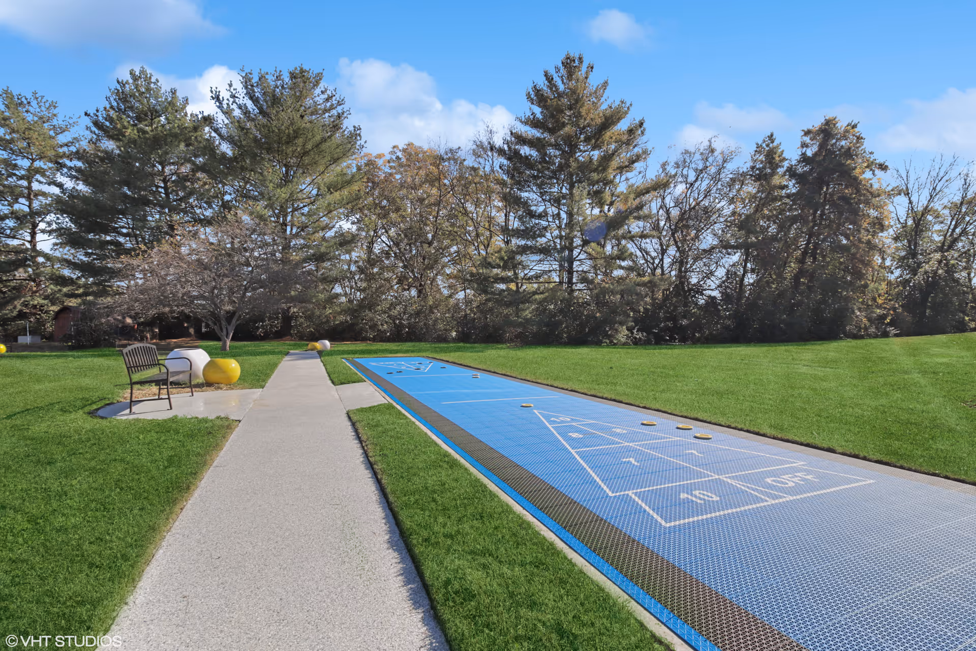 Outdoor shuffleboard court with a blue playing surface on a well-maintained green lawn. A concrete pathway runs alongside the court, with a bench and some decorative yellow and white spherical objects near the path. Tall trees and a clear blue sky are visible in the background.