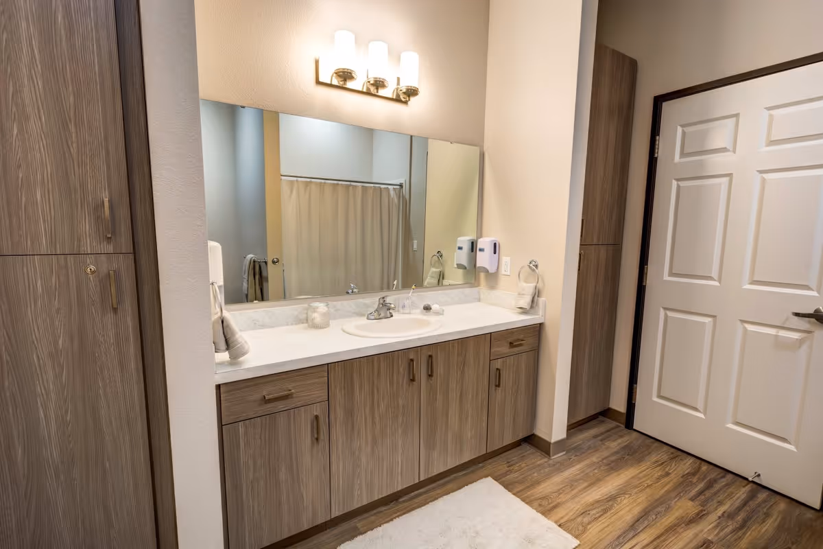A bathroom vanity area with a large mirror above a countertop with a sink. The vanity has wood-finished cabinets and drawers. Above the mirror are three modern light fixtures. On the countertop are a candle, a soap dish, and a small towel hanging on a ring. The floor has wood-like flooring and a white rug. A white door and a shower curtain are visible in the reflection of the mirror.