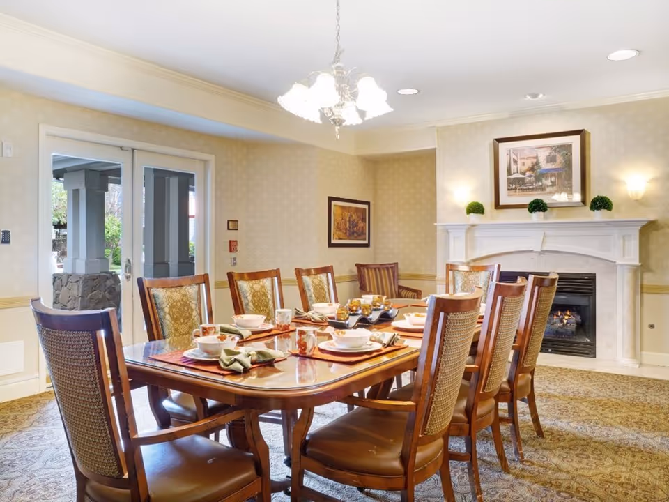 A well-lit dining room with a large wooden table set for eight people. The table is arranged with plates, bowls, cups, and napkins. The room features a fireplace with a mantel decorated with small green plants and a framed picture above it. There are two framed pictures on the walls, patterned carpet, and double glass doors leading outside.