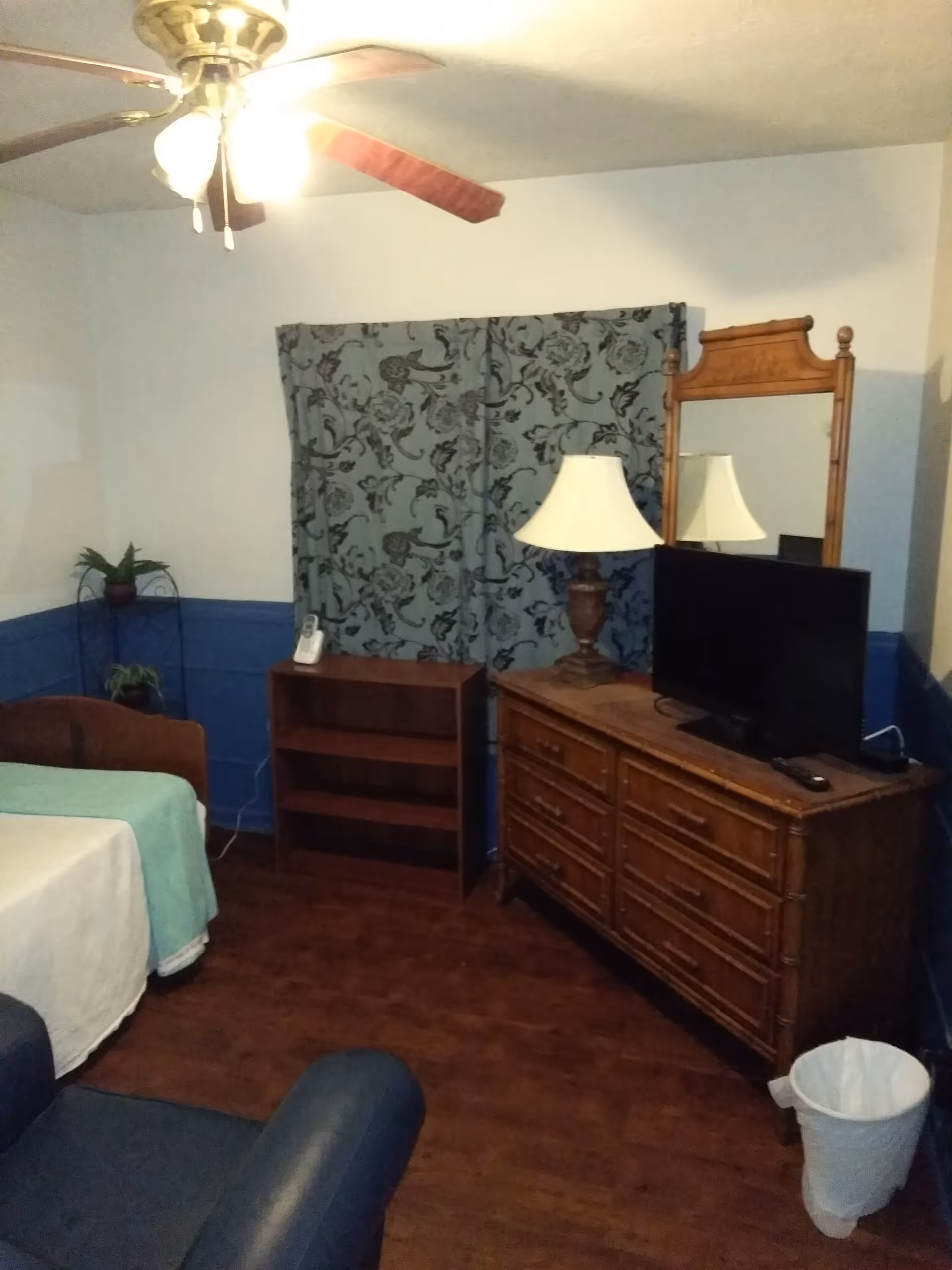 Small furnished bedroom with a bed, wooden dresser topped with a TV and lamp, a mirror, bookshelf, and a ceiling fan.
