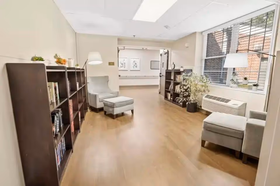 Interior senior living lounge with bookshelves, seating, plants, and a large window.