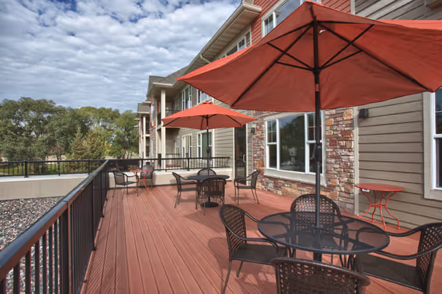 Outdoor patio area at Bel Rae Senior Living with multiple round tables and chairs under red umbrellas, adjacent to a building with stone and siding exterior walls, overlooking trees and a partly cloudy sky.