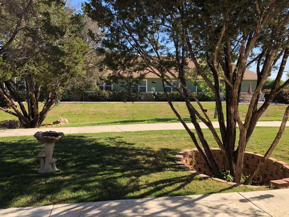 Outdoor garden area with green grass, a birdbath, and trees casting shadows. A paved walkway runs through the grassy area, and a single-story building with green siding and a brown roof is visible in the background.