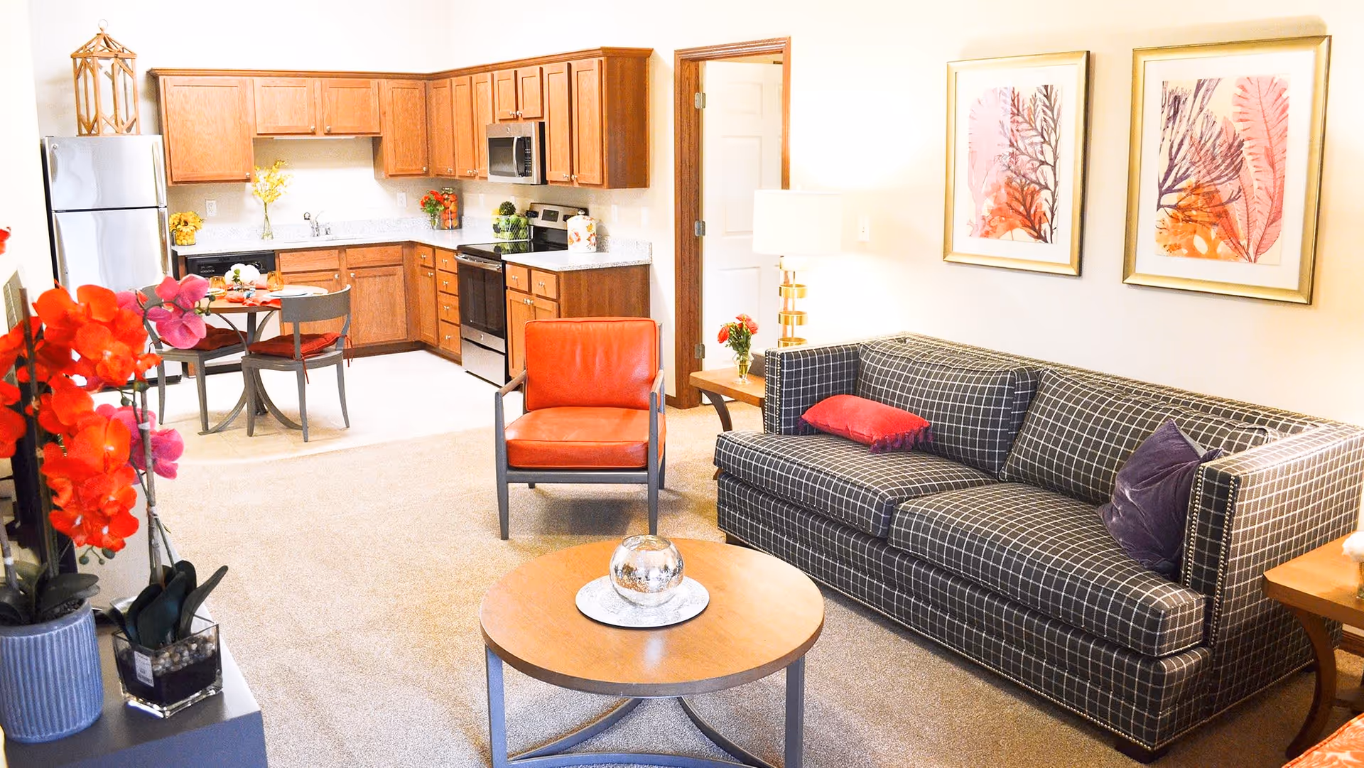 A bright and cozy living area with a checkered sofa adorned with red and purple pillows, a round wooden coffee table with a decorative glass orb, and a red armchair. The background features a kitchen with wooden cabinets, stainless steel appliances, and a small dining table with two chairs. The walls are decorated with framed artwork of colorful leaves.