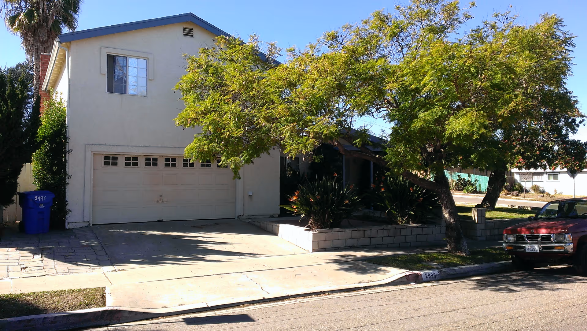 Exterior view of a two-story residential building with a white garage door, a driveway, and a large tree with green foliage in front. A red vehicle is parked on the street to the right, and a blue recycling bin is visible on the left side near the driveway.