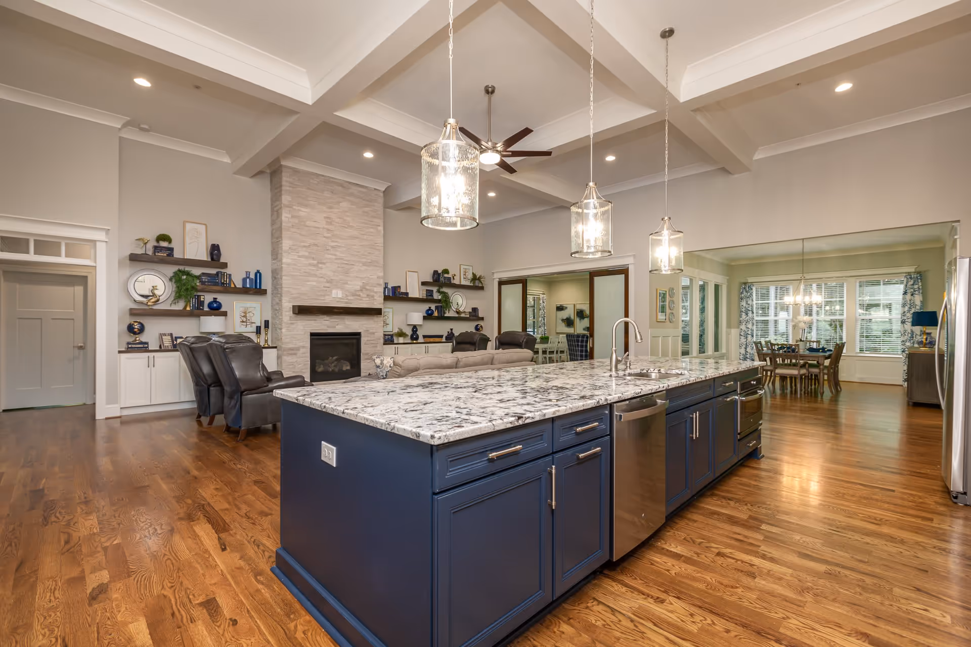 Spacious open-concept interior featuring a large kitchen island with a marble countertop and blue cabinetry, three pendant lights hanging above, a stainless steel dishwasher, and a sink. The background shows a living area with a stone fireplace, leather chairs, built-in shelves with decorative items, and a dining room with a wooden table and chairs near large windows with blue patterned curtains. The room has hardwood floors and a coffered ceiling with recessed lighting and a ceiling fan.