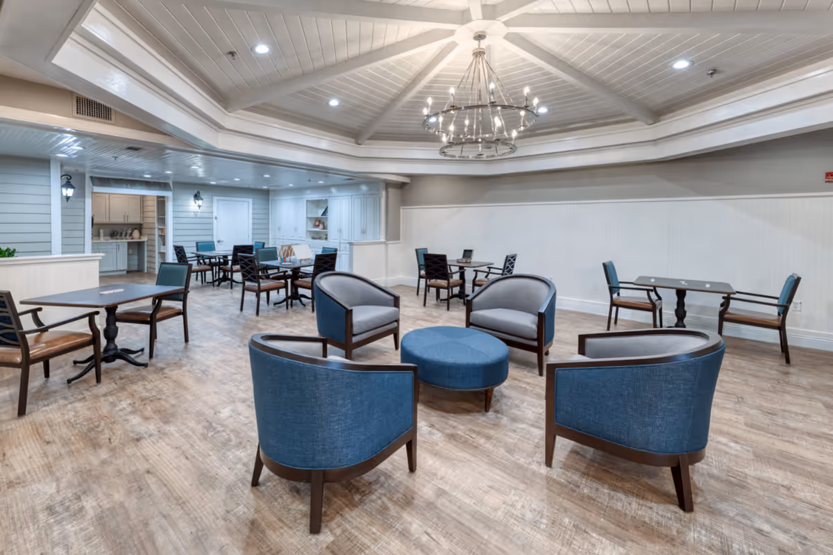 A spacious and well-lit common area with a circular arrangement of four blue and gray armchairs around a blue ottoman in the center. Several small tables with chairs are placed around the room. The ceiling features a large, elegant chandelier and recessed lighting. The floor is wood, and the walls are painted in neutral tones with white wainscoting. There is a small nook with cabinetry and shelves in the background.