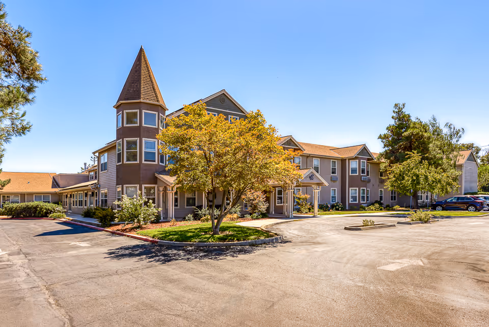 Exterior view of a senior living facility building with a distinctive turret, multiple windows, and a covered entrance. The building is surrounded by trees and landscaping, with a paved driveway and parking area in front under a clear blue sky.